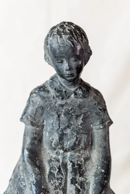 Escultura de una figura infantil de pie, con la cabeza inclinada y los brazos junto al cuerpo.